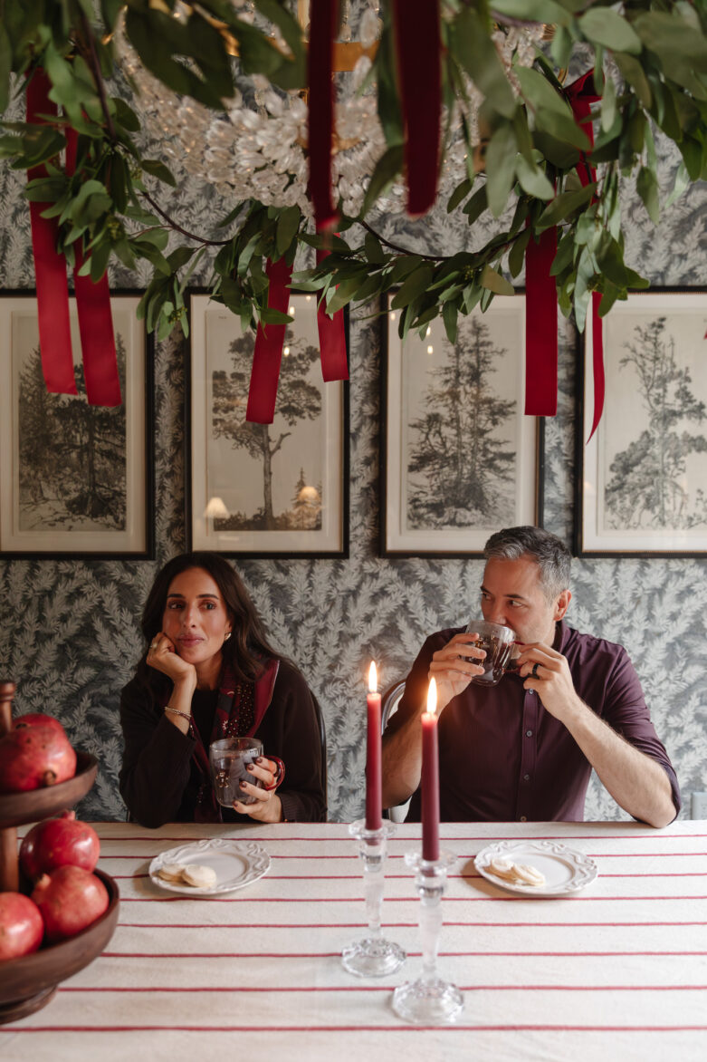 A cozy holiday dinner scene featuring a woman and man enjoying drinks at a beautifully decorated table with candles, pomegranates, and festive red accents