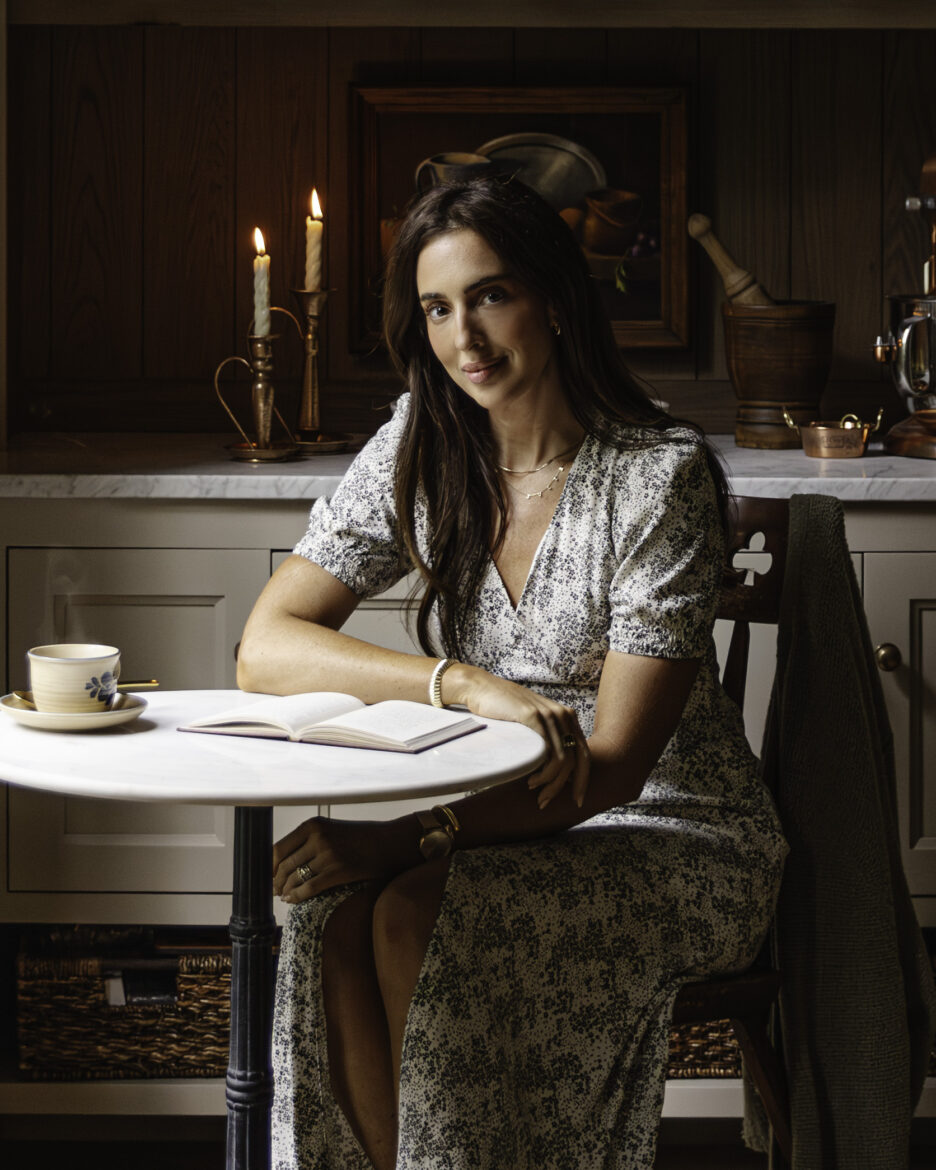 Julia sitting at the table in the kitchen in a floral dress with a mug of cider and a notepad next to her