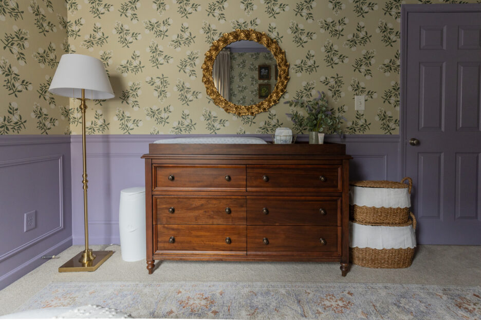 Charming room with vintage wooden dresser, floral wallpaper, gold-framed mirror, basket storage, and soft lighting for a stylish nursery or bedroom setting.