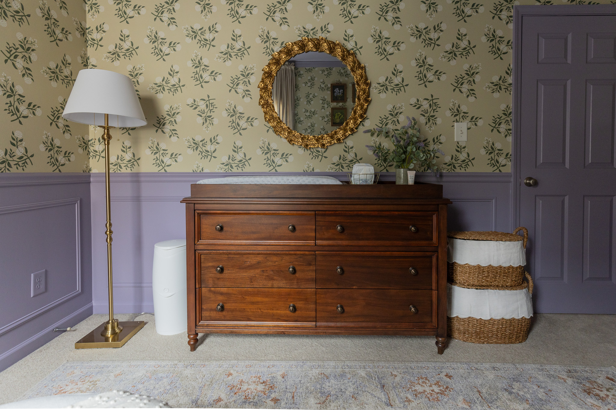 Charming room with vintage wooden dresser, floral wallpaper, gold-framed mirror, basket storage, and soft lighting for a stylish nursery or bedroom setting.