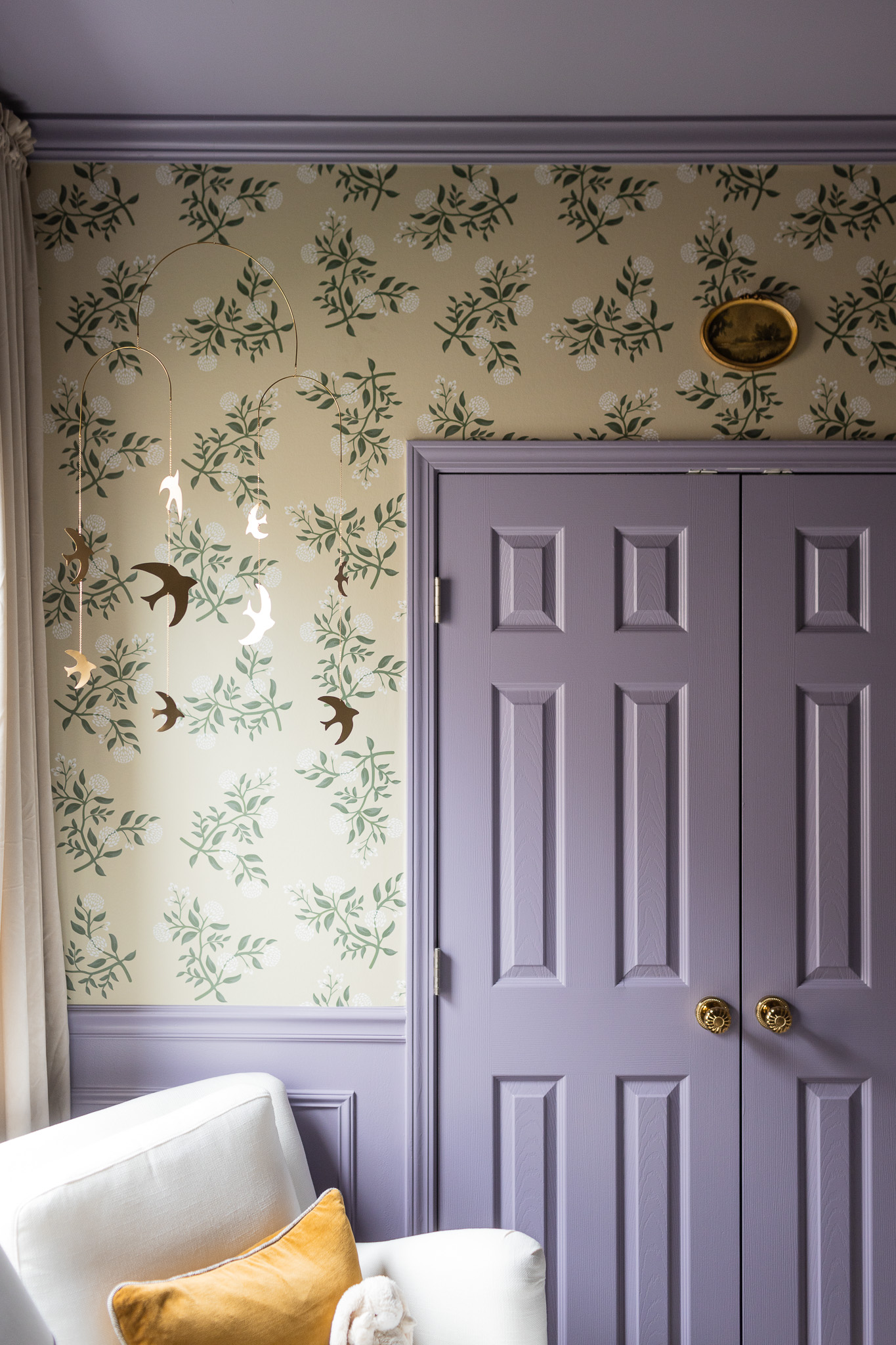 Bright nursery with floral wallpaper, purple closet doors, bird mobile, and cozy armchair for a charming kids' bedroom decor.