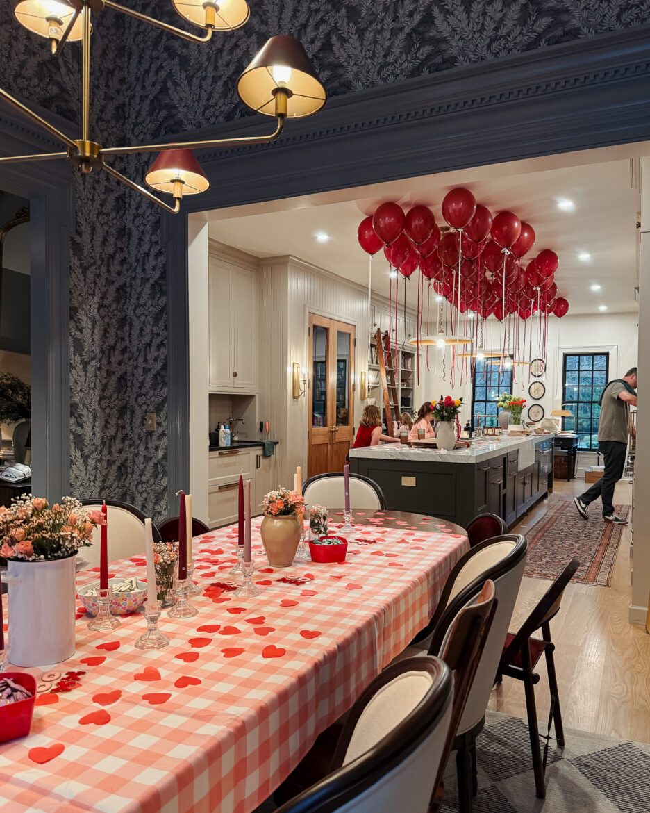 Brightly decorated dining room featuring red balloons hanging from the ceiling, a checkered tablecloth with heart-shaped confetti, floral centerpieces, and a cozy, stylish kitchen in the background.