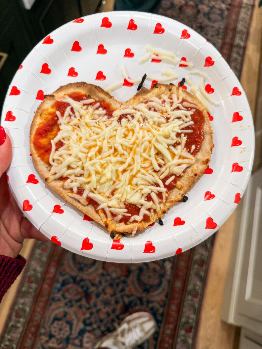A heart-shaped pizza with melted cheese and marinara sauce is presented on a decorative paper plate with red heart patterns, ideal for romantic occasions or family pizza nights.