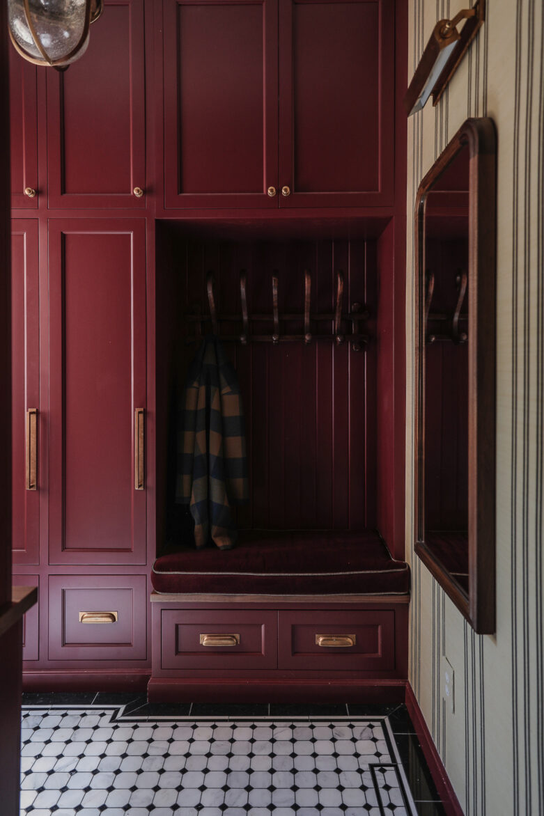 Custom burgundy mudroom with bench, hooks, gold hardware, and adjacent mirror. Perfect for organization and style.