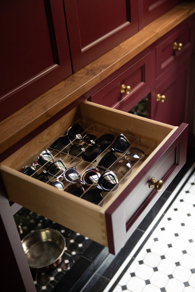 Custom built-in wooden drawer with compartments for sunglasses, in a stylish kitchen with maroon cabinets and black & white tiled flooring.