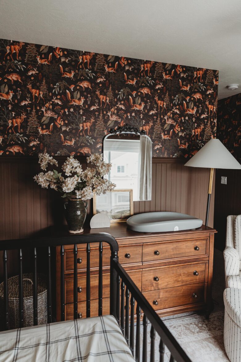 Vintage-inspired nursery decor, dark botanical wallpaper, wooden furniture, floral arrangements, and a sleek baby changing pad create a warm, inviting space for family comfort.