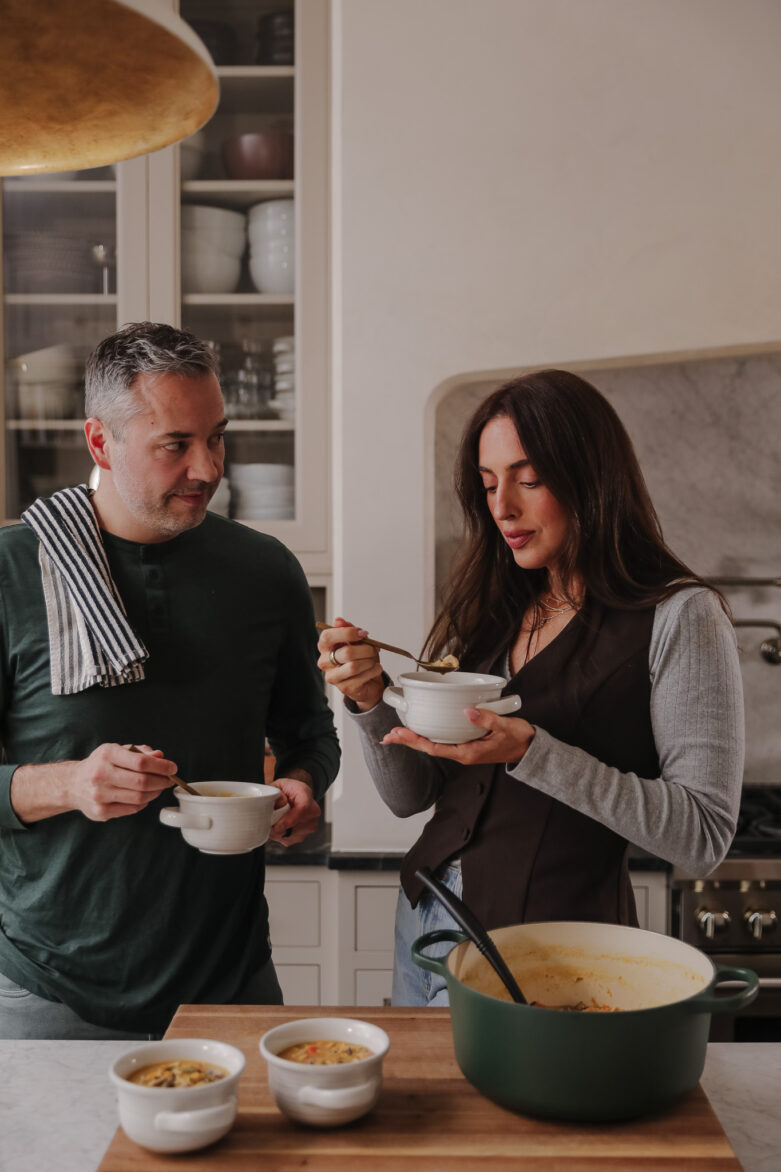 A couple in a modern kitchen, preparing a meal with bowls of soup and a large Le Creuset pot on the counter.