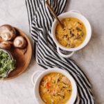 Two bowls of warm mushroom wild rice soup with fresh ingredients, served on a marble surface with a striped cloth, mushrooms, and greens nearby.