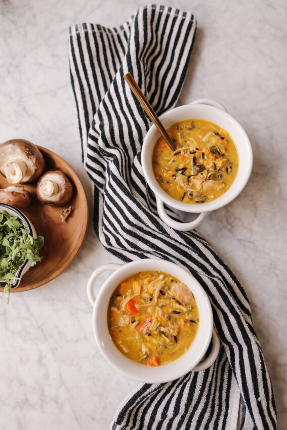 Two bowls of warm mushroom wild rice soup with fresh ingredients, served on a marble surface with a striped cloth, mushrooms, and greens nearby.