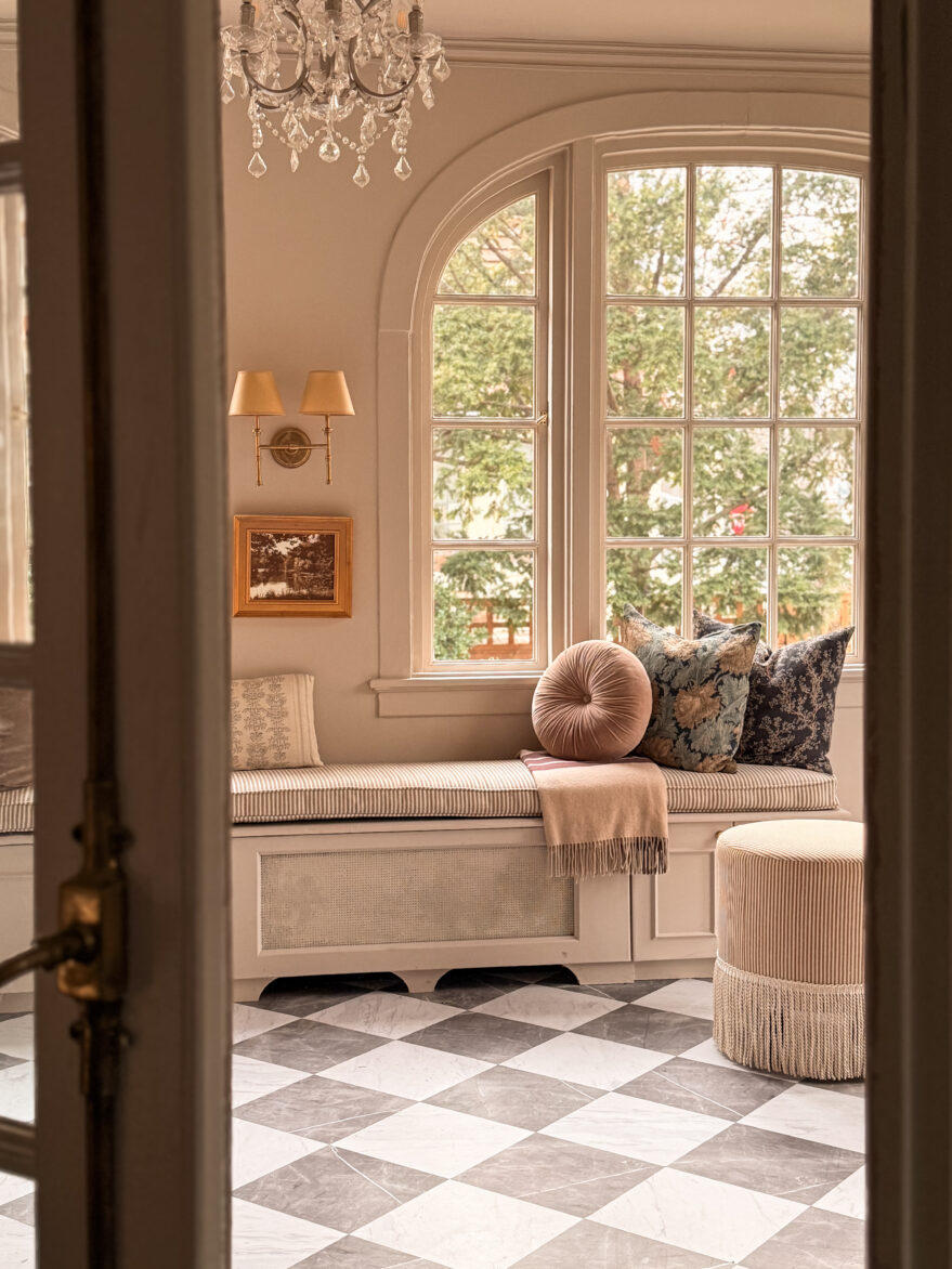 Inviting window seat with plush cushions and classic decor in a bright, airy room featuring a large arched window and vintage chandelier.