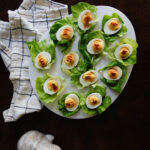 Delicious deviled eggs served on fresh green lettuce leaves, arranged on a white platter with a rustic cloth and stone heart, perfect for healthy appetizers and family gatherings.