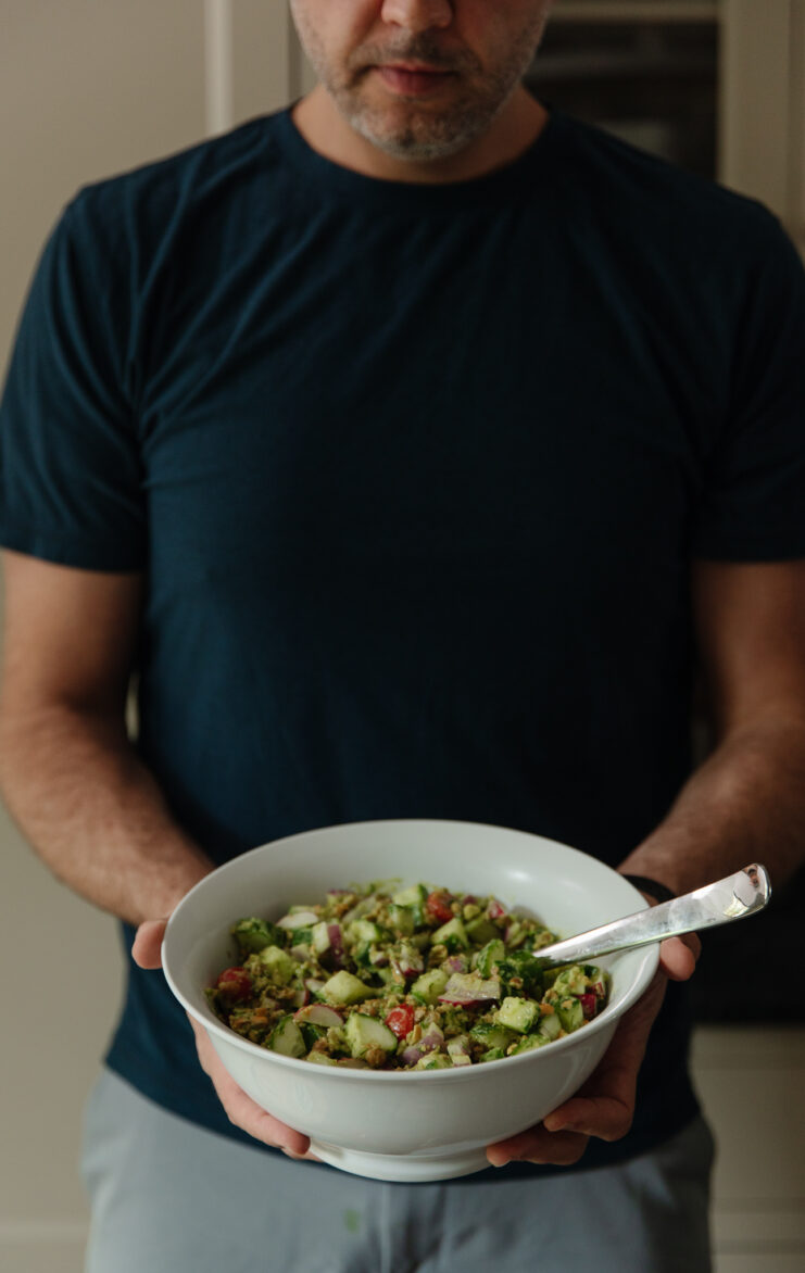 Close-up of Chris holding a large white bowl of colorful vegetable salad including cucumber, tomato, radish, and avocado, in a modern kitchen setting.
