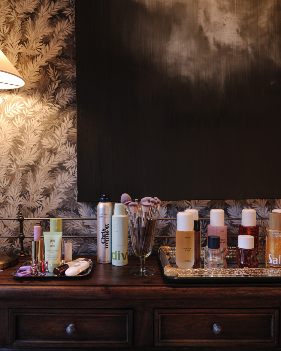 Stylish vanity setup featuring skincare, makeup, and hair products arranged on a dark wooden dresser against a black wall and patterned wallpaper.