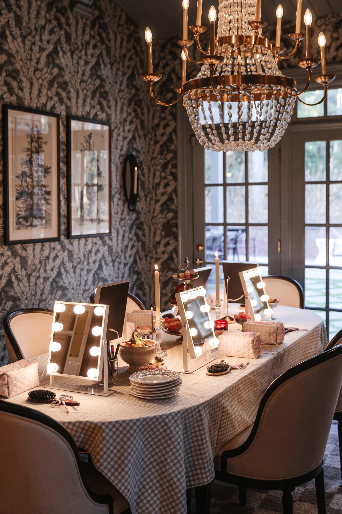Chandelier and vanity mirrors illuminate a stylish dining table set for a celebration, with elegant decor and natural light streaming through large windows in a sophisticated dining room.