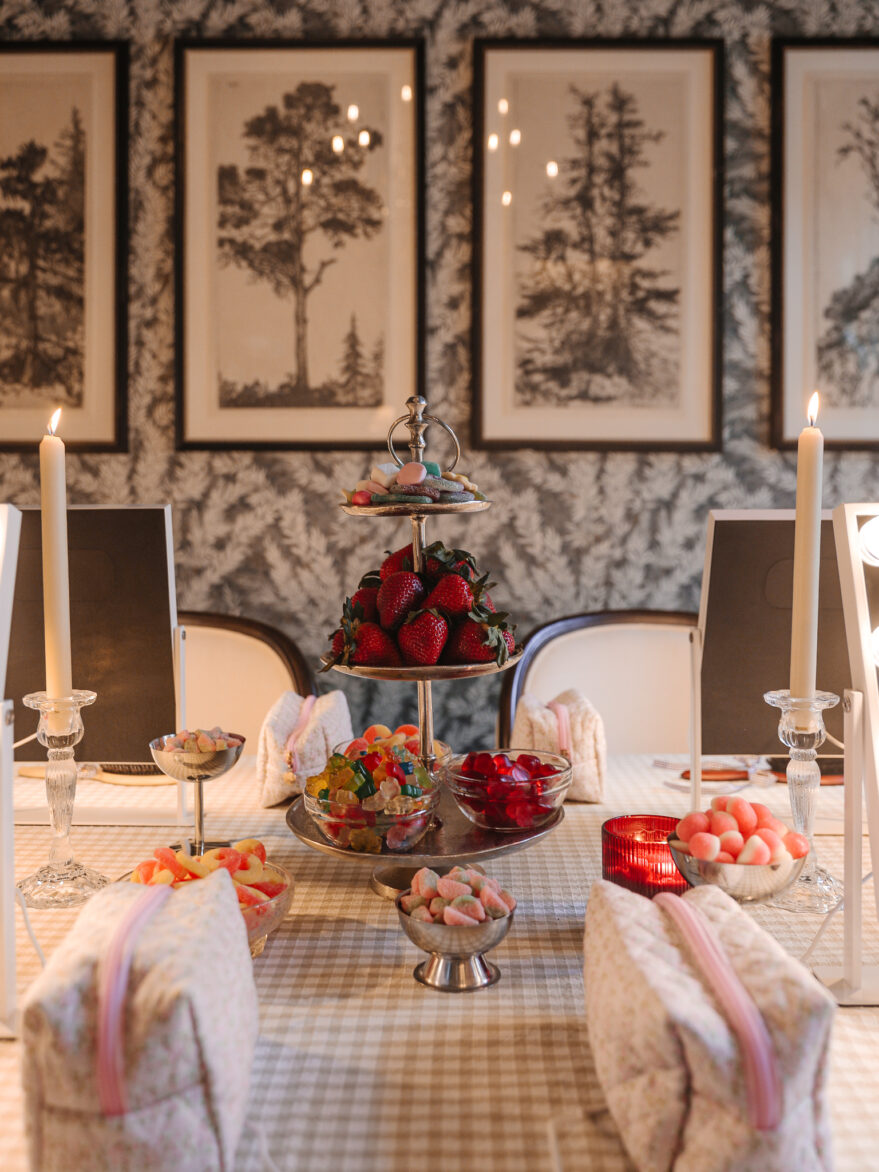 A beautifully decorated dining room featuring a table set with colorful candies, strawberries, and pink marshmallows, surrounded by white chairs with pink accents, and framed black-and-white nature ar.