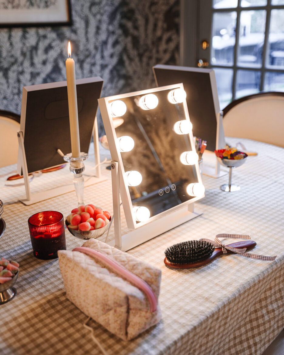 Lit makeup vanity featuring a mirror with surrounding bulbs, a candle, and various beauty tools on a checkered tablecloth.