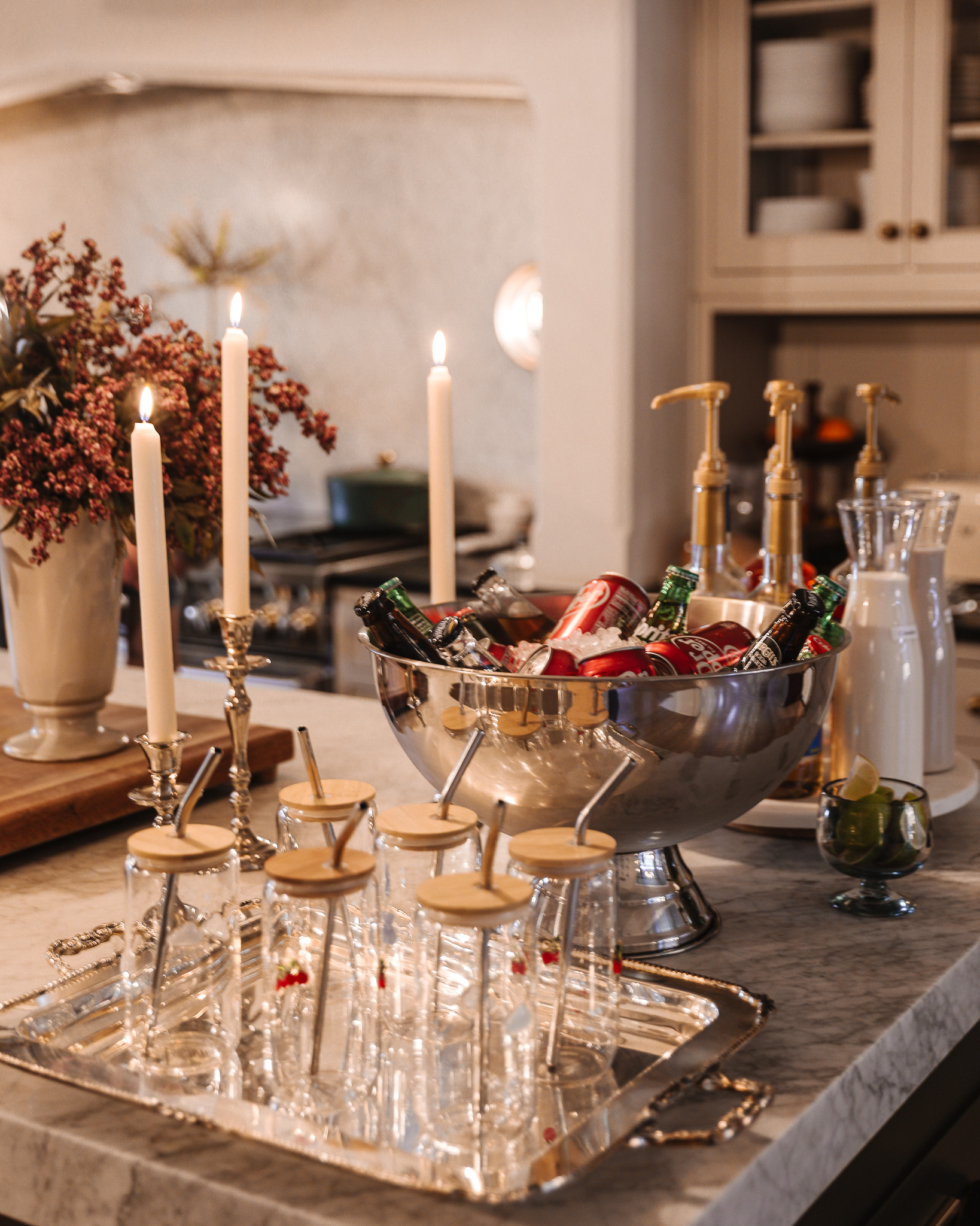 A beautifully decorated kitchen island with a large silver bowl filled with holiday drinks, surrounded by lit candles, glass jars with straws, and festive decor for a cozy celebration.