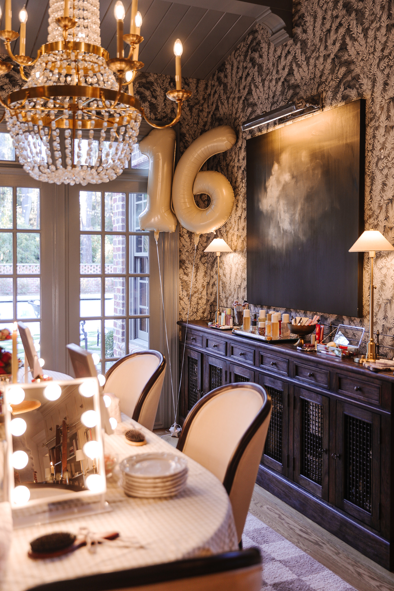 A stylish dining area featuring a crystal chandelier, balloon numbers "16," and a dark wooden sideboard with decorative items, creating a festive and sophisticated atmosphere.