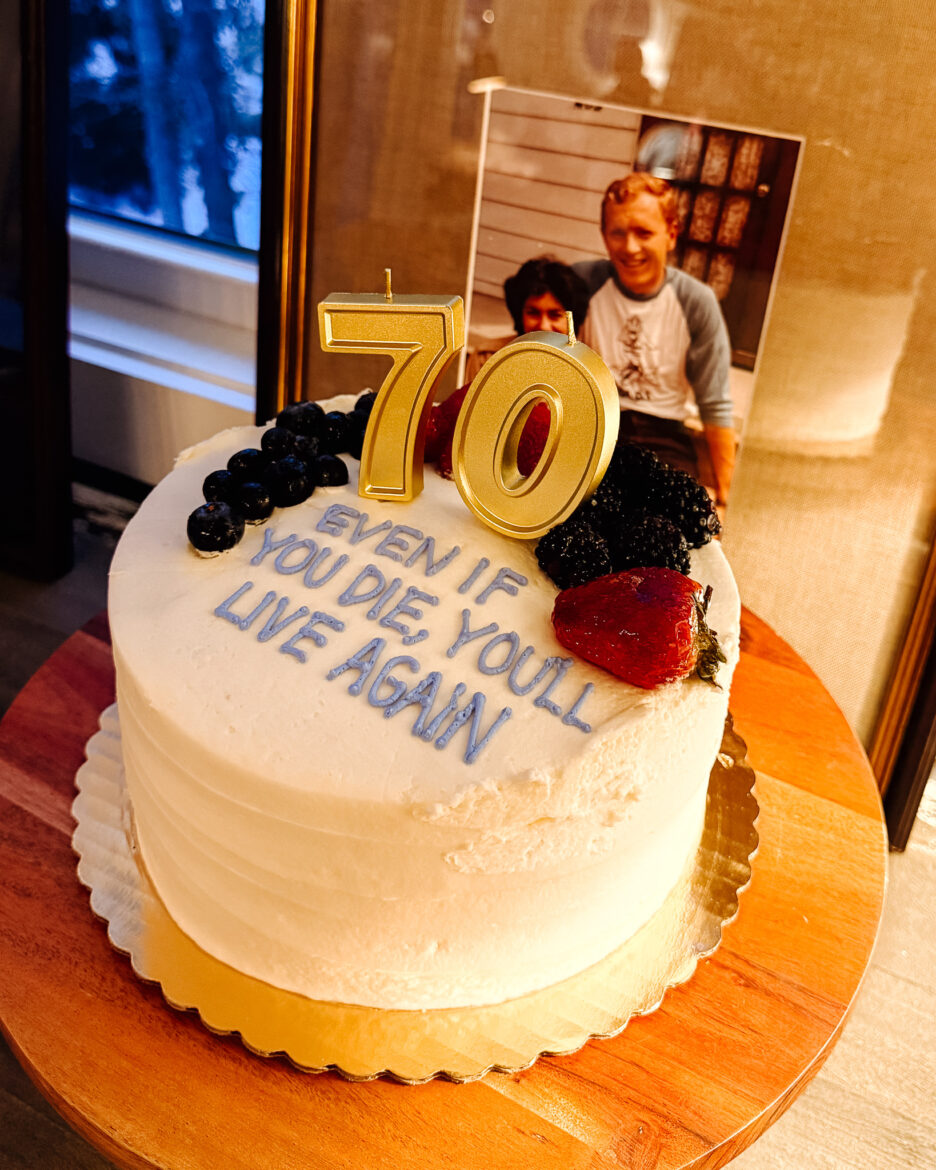A birthday cake decorated with a photo of two children, a large gold "70" candle, and fresh berries, celebrating a 70th birthday milestone.