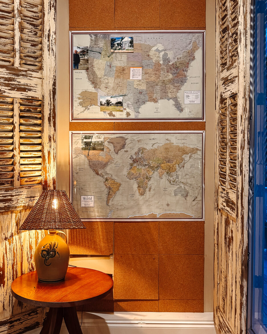 A detailed map of the United States and the world displayed on a wall, framed and mounted in a cozy, rustic room with wooden shutters and warm lighting.