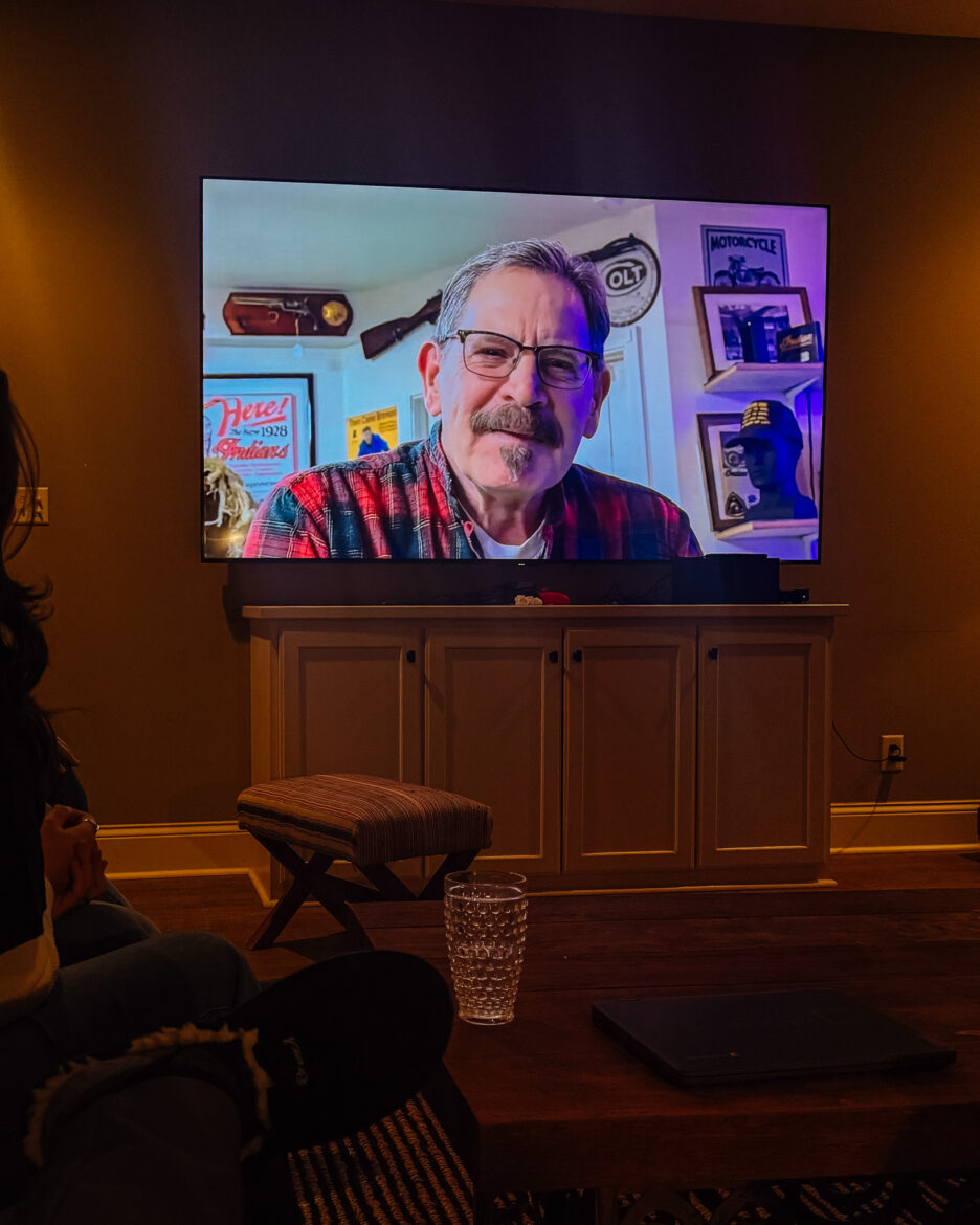 Man on a video call displayed on a large TV screen, wearing glasses and a plaid shirt, in a cozy home office with vintage decor and collectibles on the shelves.