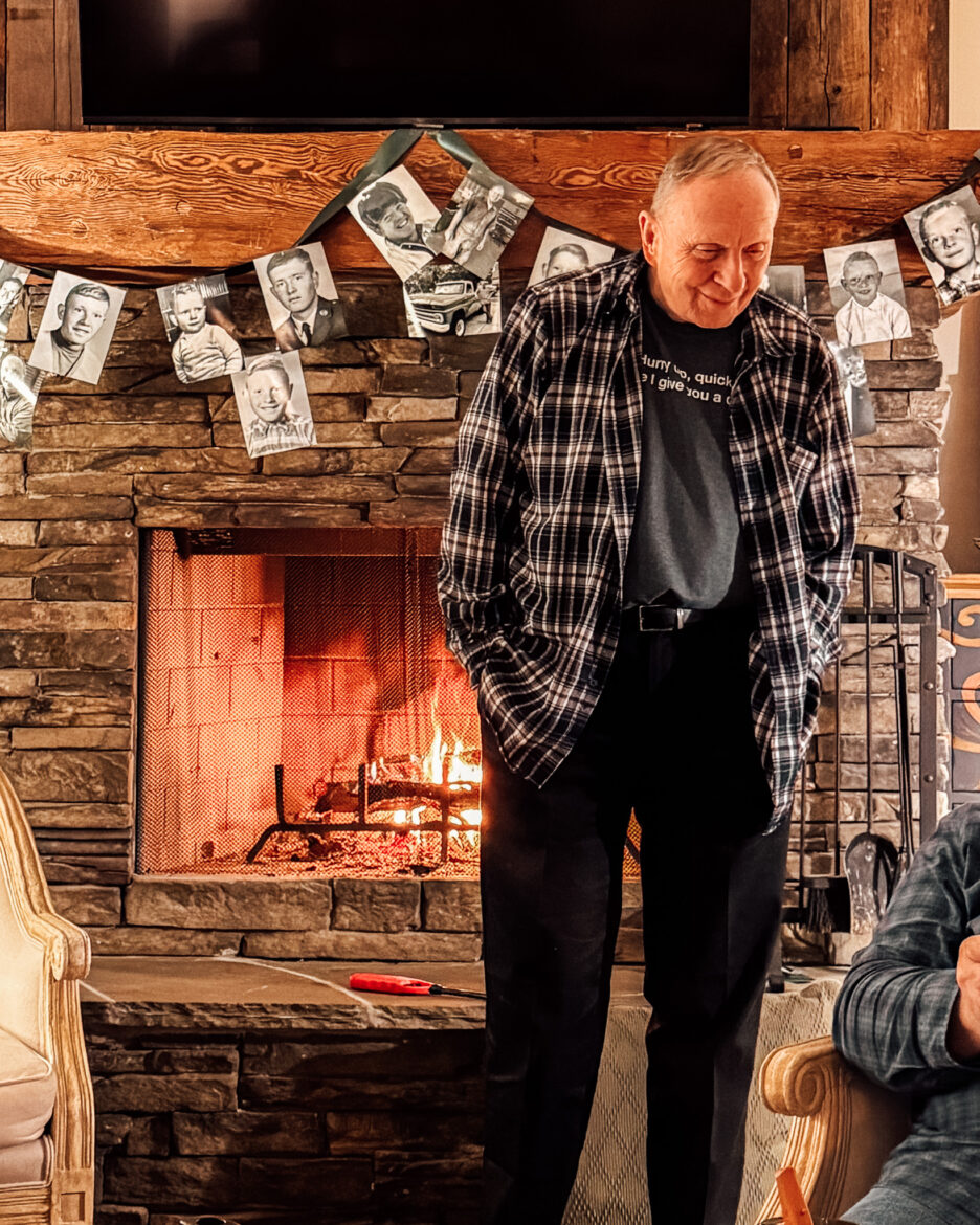 Man standing near a lit fireplace in a cozy living room with family photos hanging above the mantel.