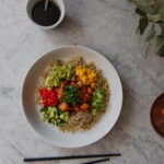 Healthy Asian rice bowl with vegetables, mango, cucumber, and salmon, garnished with green onions and served with Asian-style sauce.