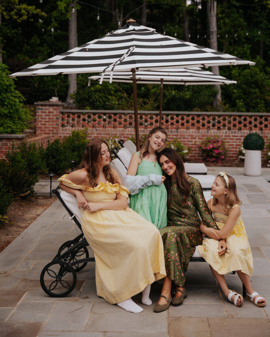 Mother and daughters relaxing outdoors with children under a large striped umbrella.