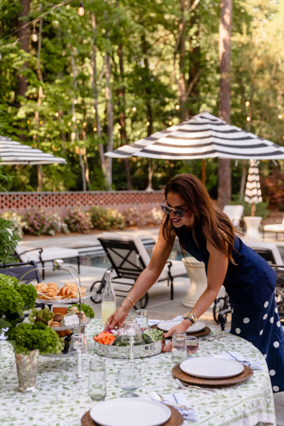 Woman setting up an outdoor garden party table with floral decorations, glassware, and tableware in a lush, green backyard with string lights and umbrellas.