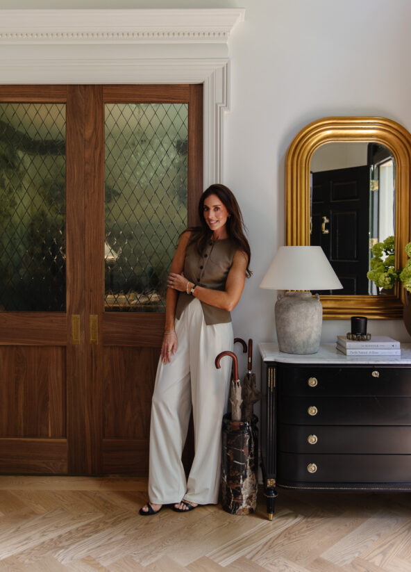Julia Marcum standing in a stylish home entryway with wooden double doors, a black dresser, a white table lamp, and a gold-framed mirror, showcasing modern interior design and cozy decor.