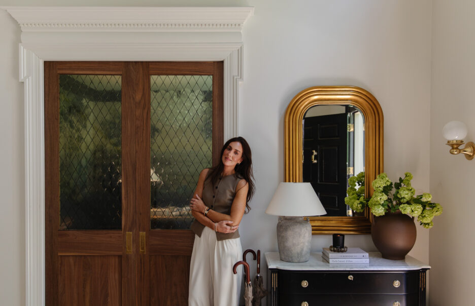Julia Marcum standing in a stylish entryway with wooden double doors, a gold-framed mirror, and a black console table with a white lamp and green floral arrangement.