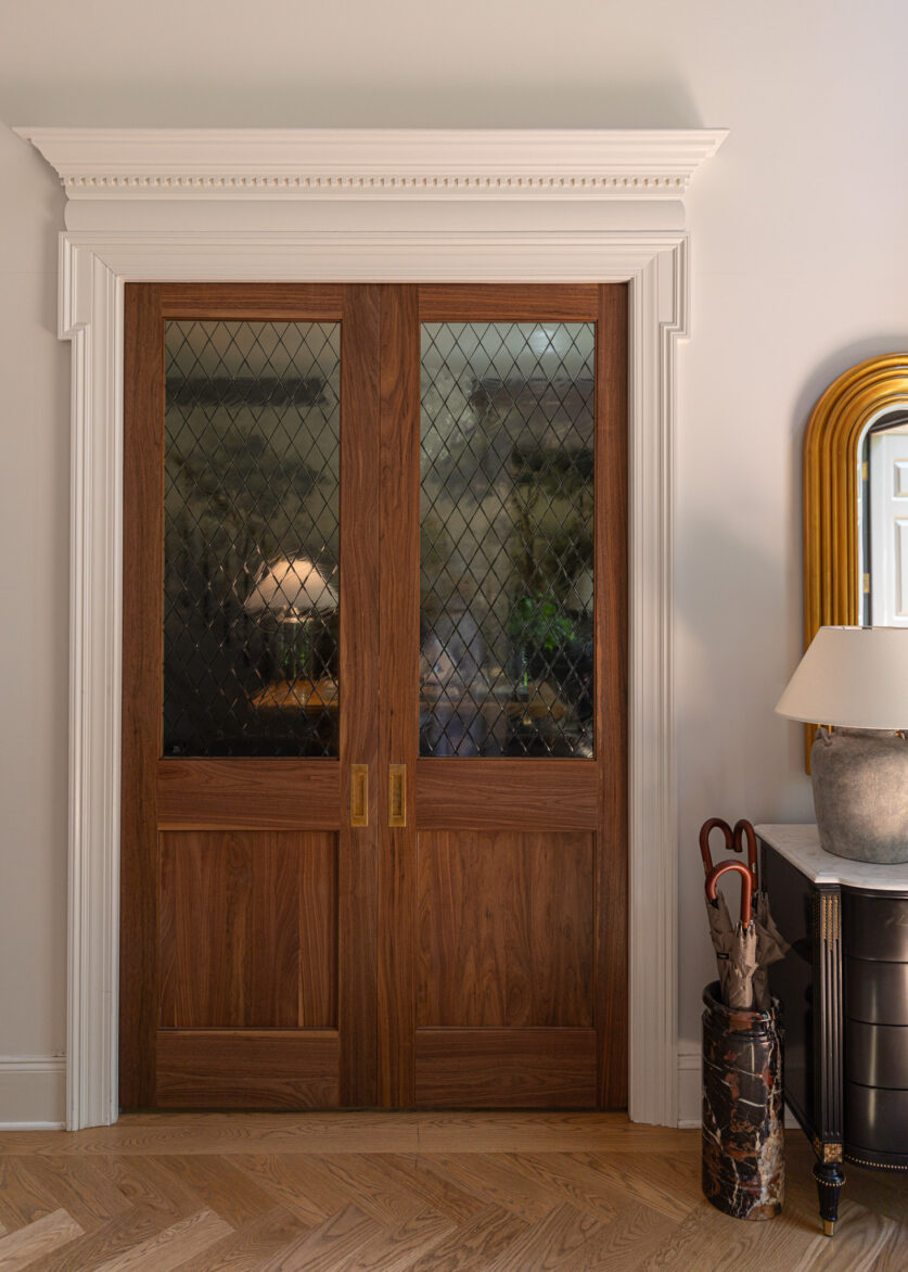 Charming interior space showcasing wooden pocket doors with diamond-patterned window film on glass, complemented by a black dresser, a large decorative mirror with a gold frame, and a table lamp. Ideal for stylish home design inspiration.