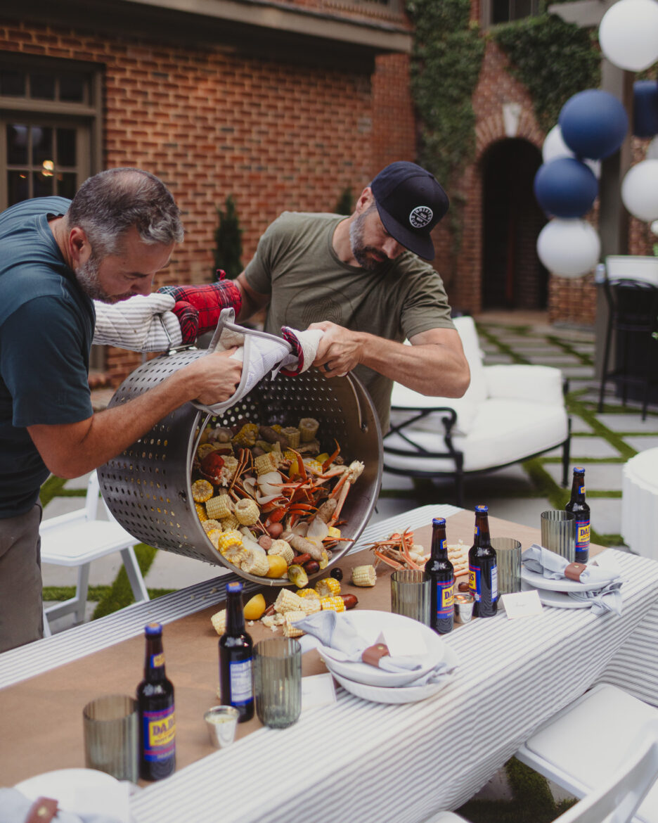 Two men are preparing a large seafood boil outdoors, pouring cooked seafood and vegetables from a large metal pot onto a table set for a casual meal. The scene features a brick building, string lights, and a relaxed, friendly atmosphere.