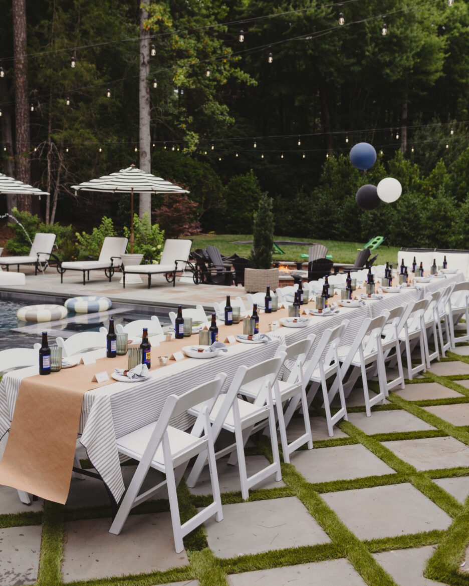 Outdoor dinner party with a long table, white chairs, and string lights in a lush backyard setting. Ideal for summer celebrations and outdoor entertaining.