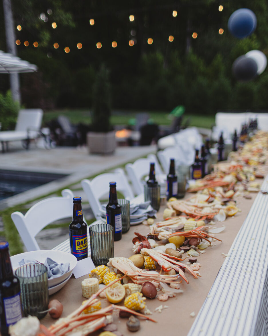 A backyard dinner party scene featuring a long table with food and drinks, white chairs, and decorative balloons, set against a brick house and greenery.