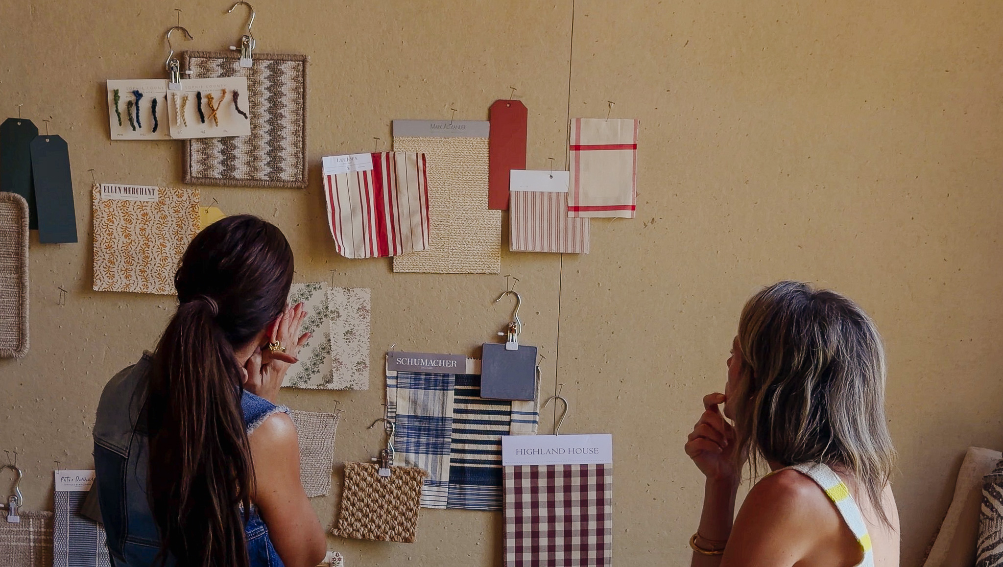 Two women examining fabric swatches and textile samples on a wall for interior design inspiration.