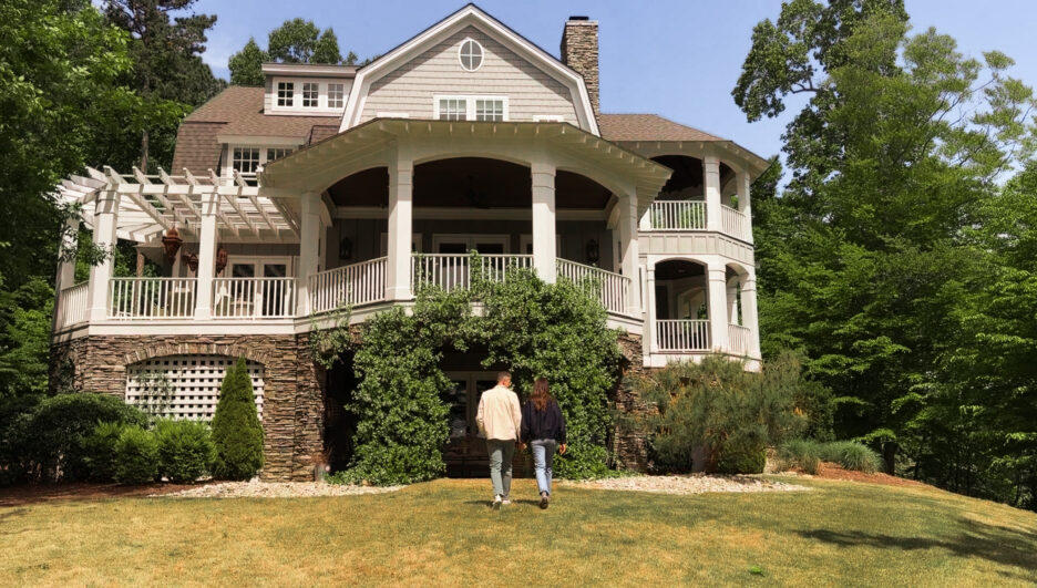 Beautiful Craftsman-style house featuring a spacious wraparound porch, white railings, and lush greenery, set against a backdrop of tall trees and a clear blue sky.