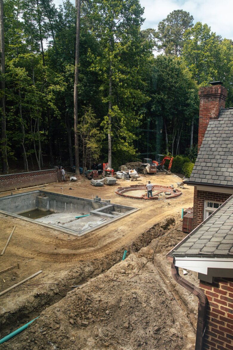 Construction site for a backyard pool with surrounding dirt, construction equipment, and workers, with a wooded background and a brick house.