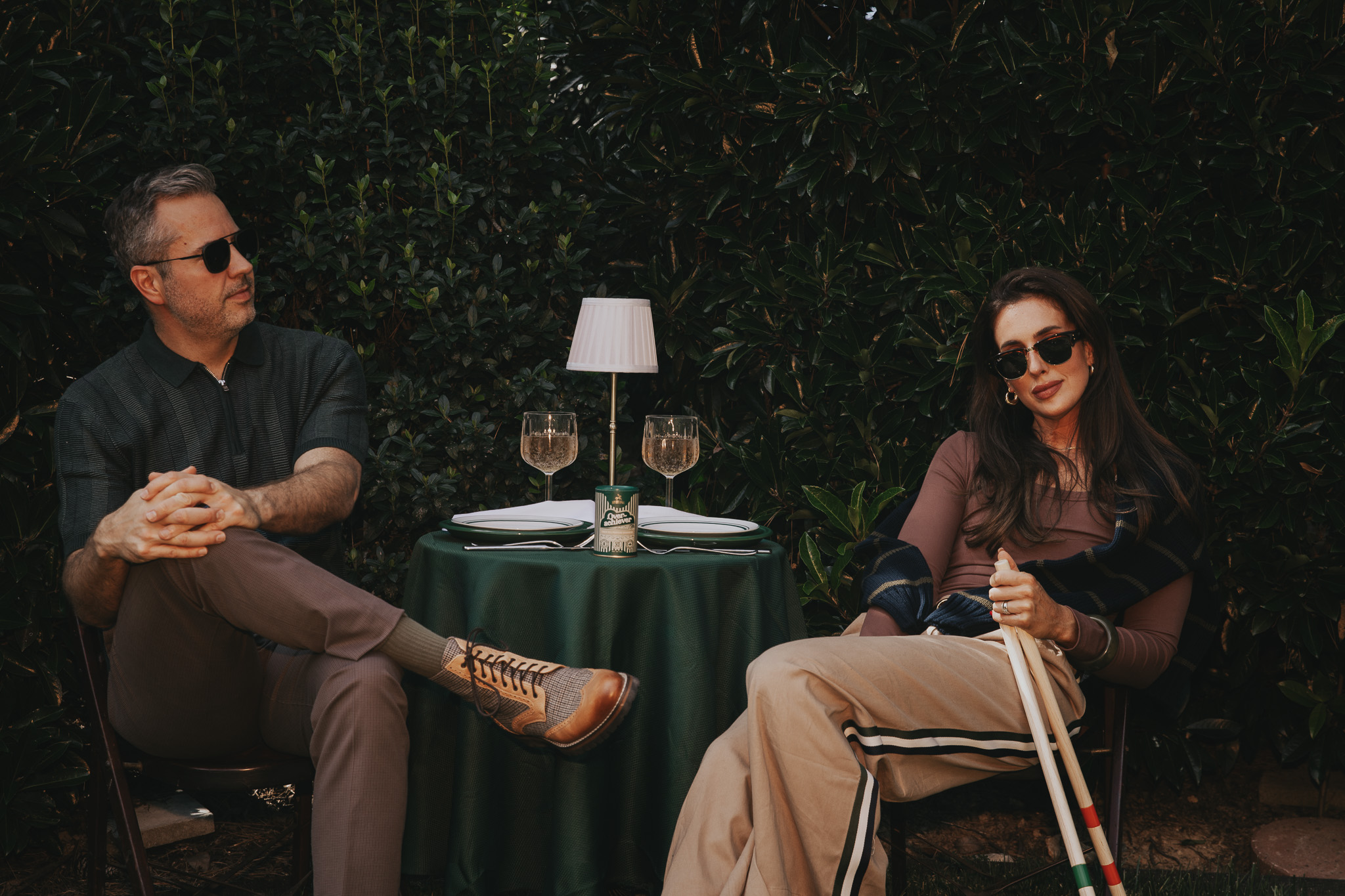 Chris and Julia seated at a small green cafe table set for dinner outdoors, a can of Hey Old Sport Overachiever seasoning at the center.