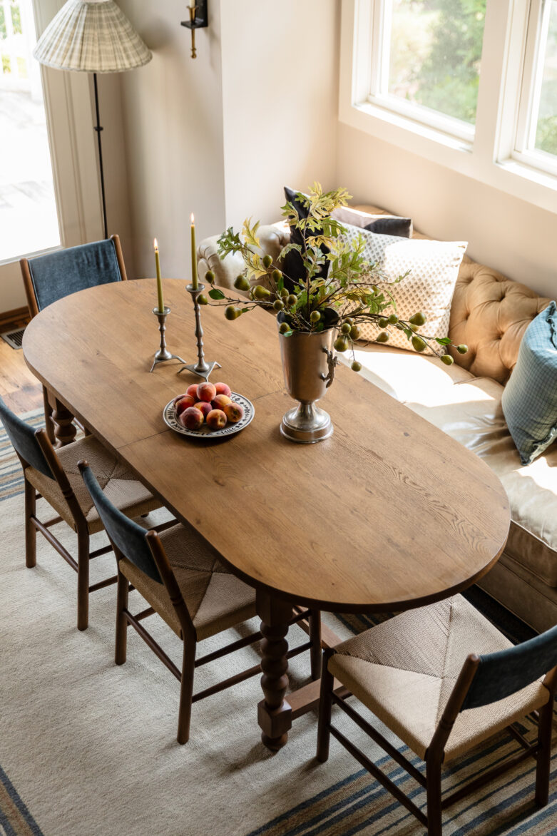 A high-angle shot of a warm traditional dining space showing an oval wood table, woven dining chairs, and a tufted built-in banquette booth beneath bright windows.