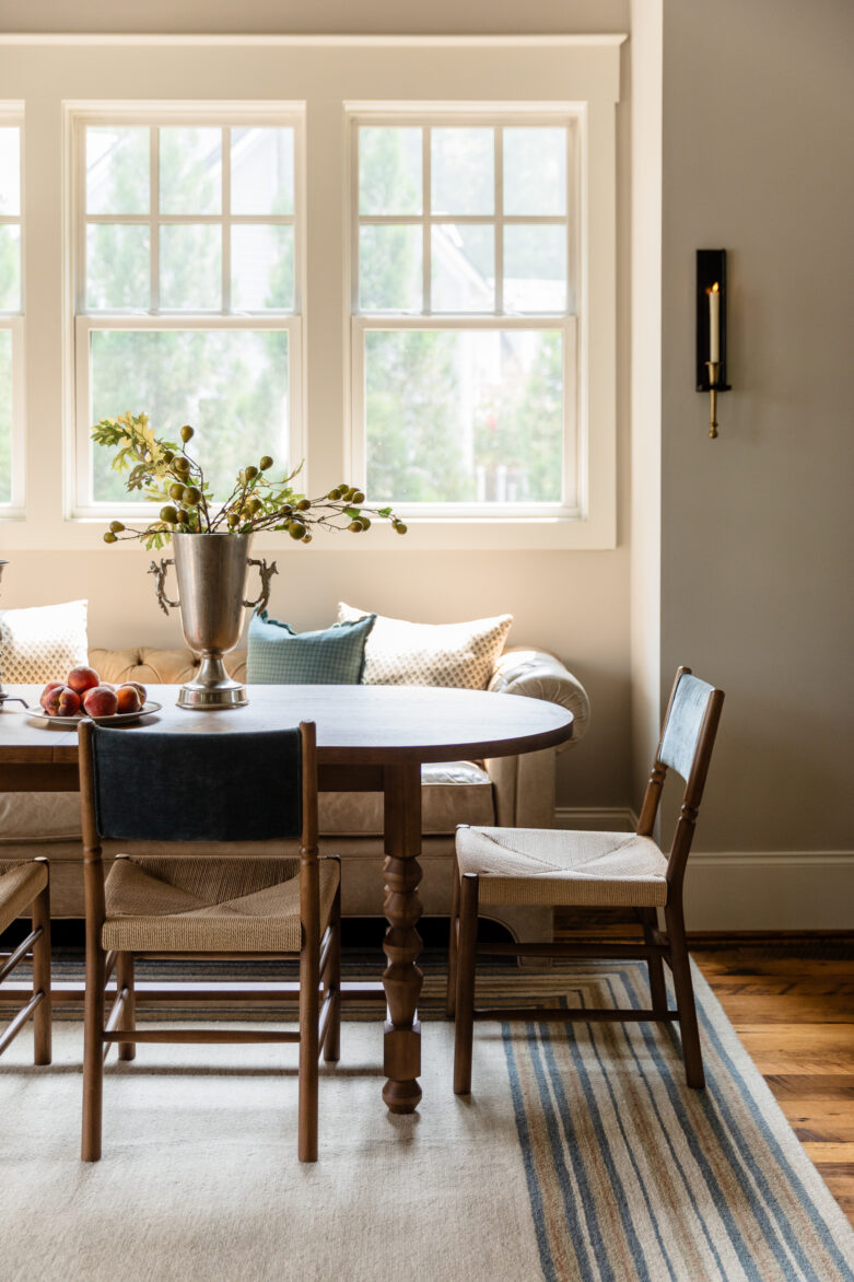 A warm, modern traditional dining space with a wooden table, woven chairs, a tufted sofa booth, and a striped area rug by a bright window.