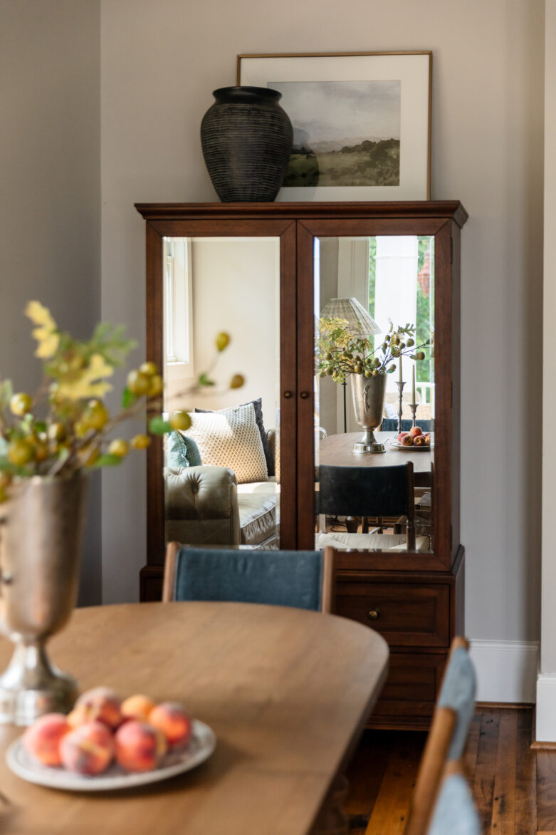 Modern traditional dining room styling showcasing a rich wood armoire with mirrors reflecting the room, styled with vintage-inspired tabletop decor, a textured black vase, and a gold-framed landscape painting.