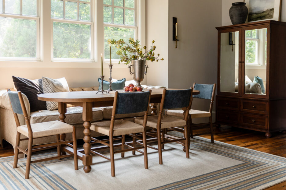 Transitional dining room styling featuring a warm wood table, woven wood chairs, a cream tufted banquette, a striped rug, and a modern black candle sconce on a neutral wall.