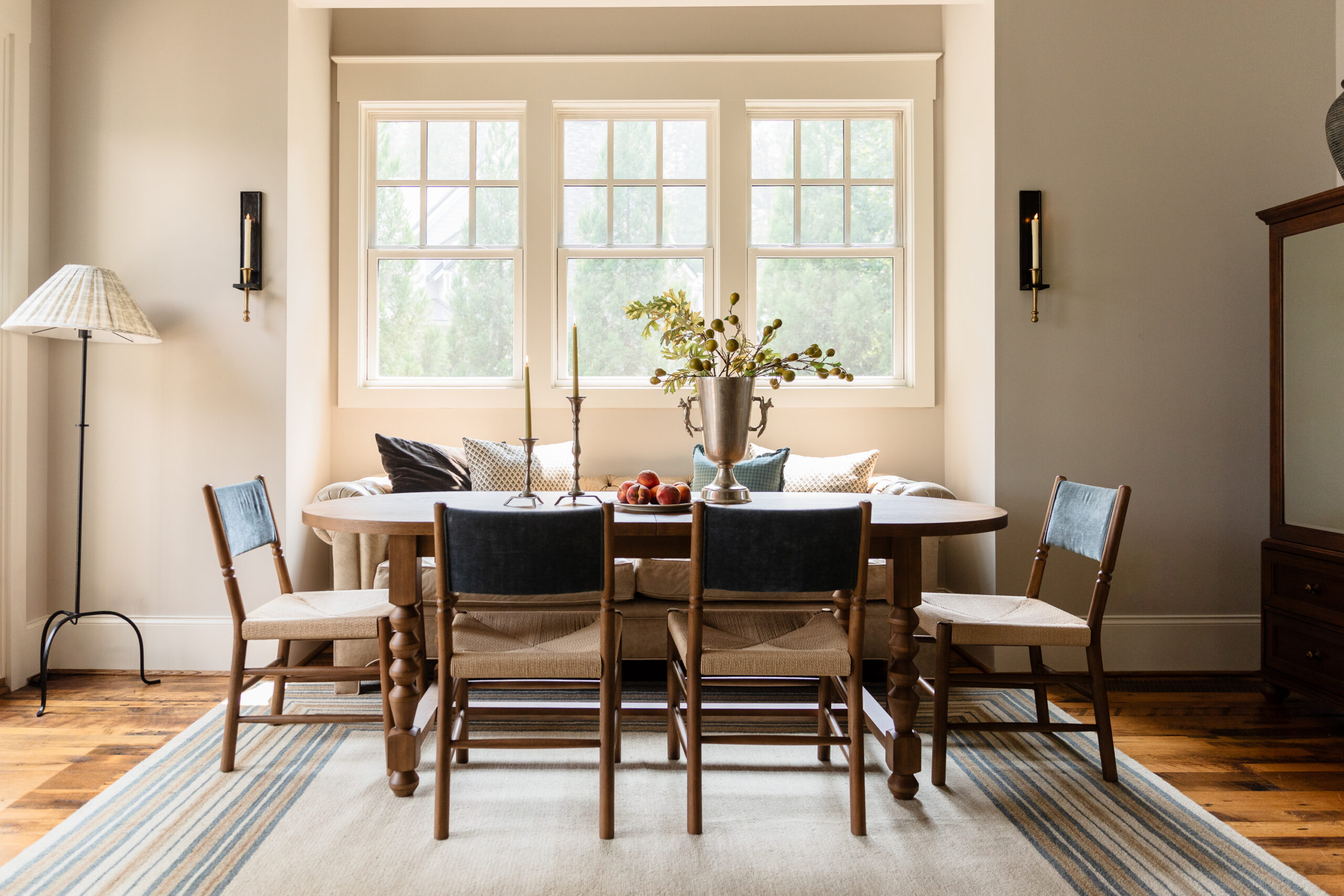 Transitional dining room styling featuring a warm wood table, woven wood chairs, a cream tufted banquette, a striped rug, and a modern black candle sconce on a neutral wall.