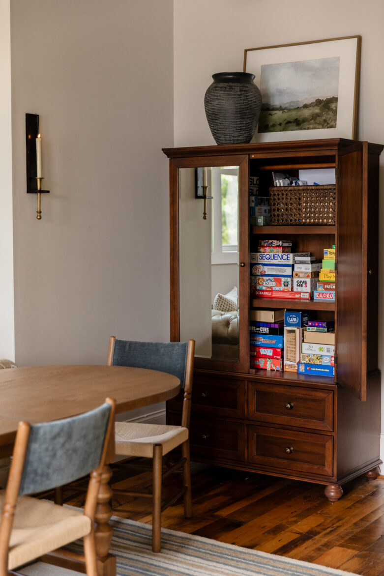 Modern traditional dining room styling showcasing a rich wood armoire with mirrors reflecting the room, styled with vintage-inspired tabletop decor, a textured black vase, and a gold-framed landscape painting and board games inside